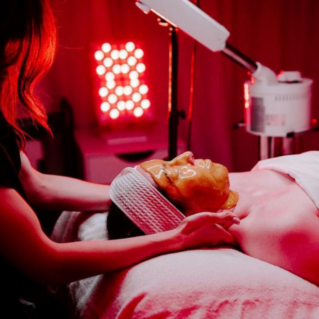 One woman providing skincare to another woman laying down with a red light therapy device in the room.
