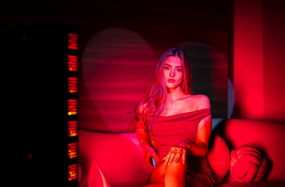 Woman sitting beside a red light therapy panel at home, the kind used for red light therapy for face and full-body treatment