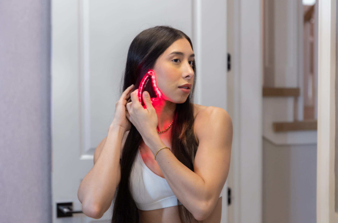 A young girl in a bathroom using Vital Red Light's red light therapy gua sha on her face.