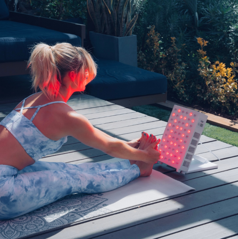 A woman stretching her legs in front of Vital Red Light's Vital Pro in front of her feet, showing the difference of red light therapy vs microcurrent.