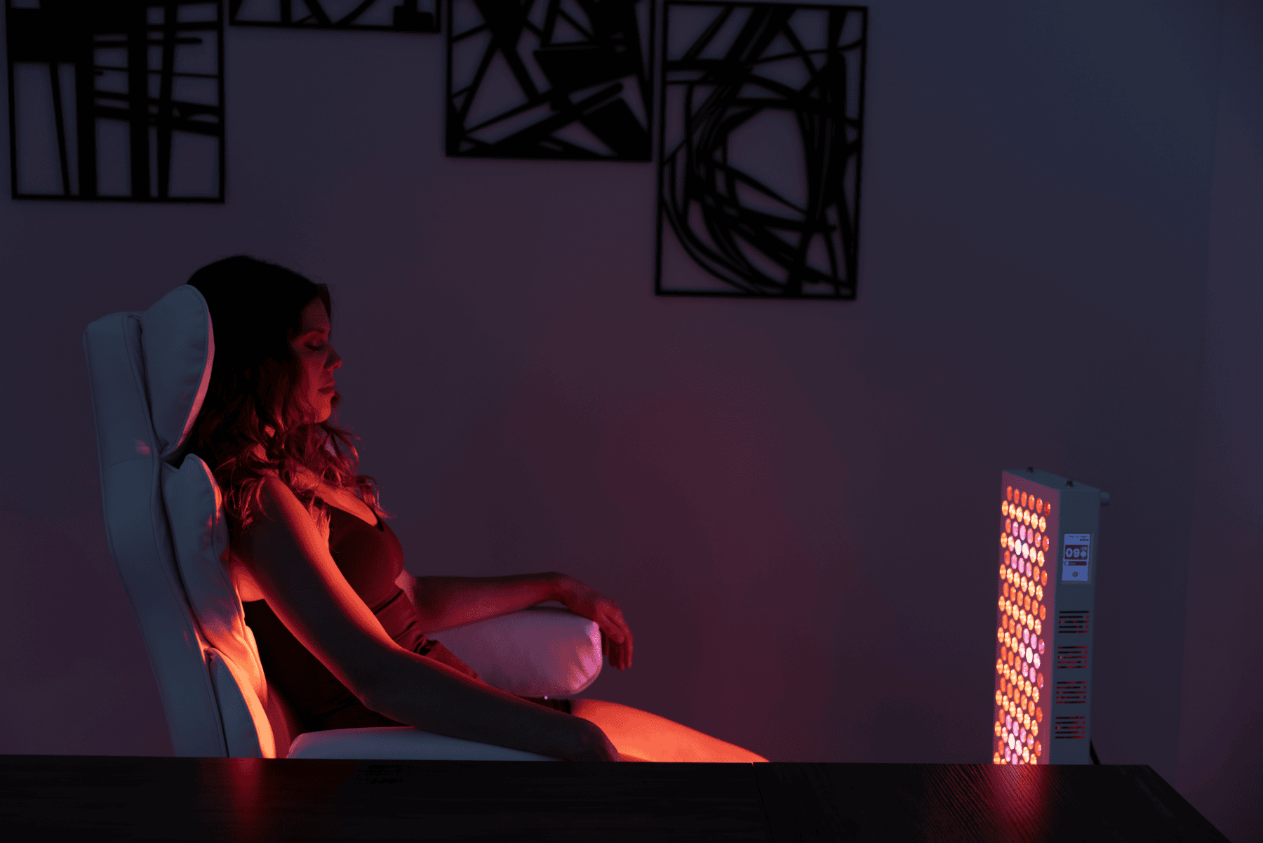 A woman sits in a chair facing the Vital Pro 2.0 light therapy panel, which emits red and yellow light in a dimly lit room adorned with abstract wall art.
