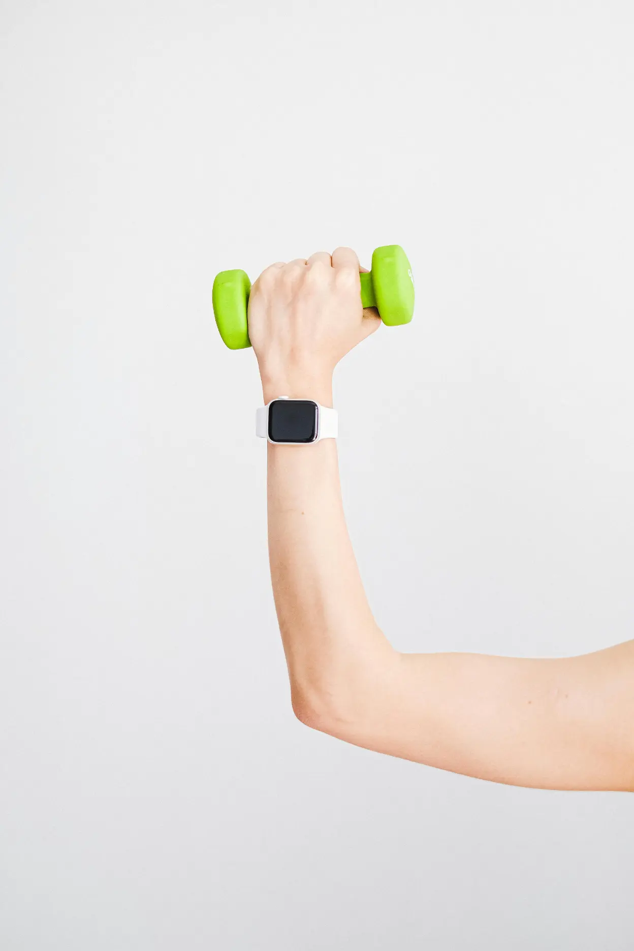A hand wearing a smartwatch holds a small green dumbbell against a plain white background biohacking health