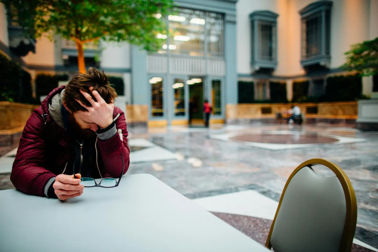 Person sitting at a table in a spacious indoor area, holding glasses and resting their head in one hand, clearly showing the strain as stress impacts the brain. A few other people are in the background, subtly adding to the contemplative atmosphere.